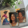 Three gratitude journals displayed on a glass table next to a plant. The journals have distinct cover designs featuring women in various contemplative poses.